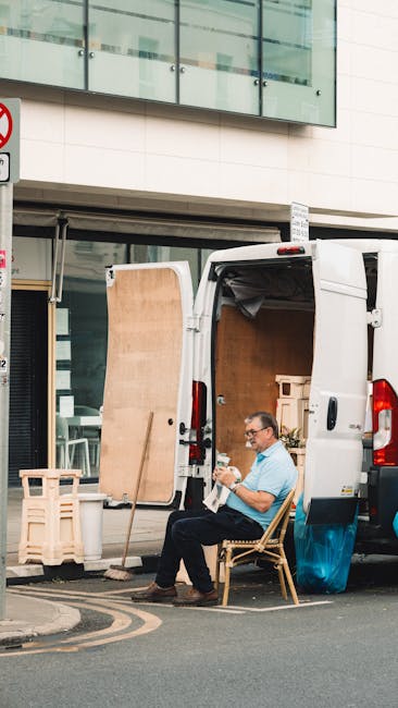 A man wearing glasses and a light blue shirt is seated on a wooden chair next to the open rear doors of a white cargo van, which is parked on an urban street in front of a modern building with glass windows. Inside the van, visible wooden panels and cardboard boxes are part of the packing materials used in a home relocation or furniture transport process. The man appears to be reading a document or a mobile device, possibly taking a break during a house removal or moving service provided by Man with Van Acton. The scene includes a pavement area with additional packing supplies, such as stacked wooden crates or pallets, nearby. Surrounding elements include a traffic sign and pavement markings, indicating a street loading zone or parking area, with natural daylight illuminating the scene and emphasizing the logistics involved in professional removals and packing operations.