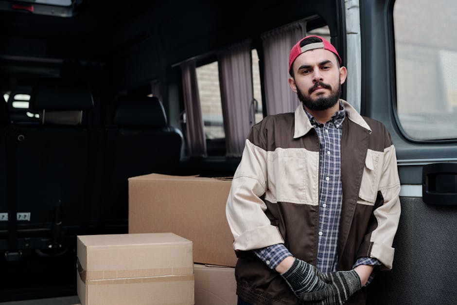 A man with a beard, wearing a red cap backwards, a checkered shirt, and a two-tone work jacket, stands beside cardboard moving boxes inside the back of a van. The van has dark interior with visible seats and a window framed by light-colored curtains. The man appears to be involved in a home relocation or furniture transport process, suggesting a professional removals service with Man with Van Acton. The cardboard boxes are sealed with packing tape, indicating they are prepared for moving or storage, and are placed on the pavement or loading area next to the van. The lighting is natural, and the focus is on the individual and the packing materials, emphasizing a routine step in packing and loading during house removals.