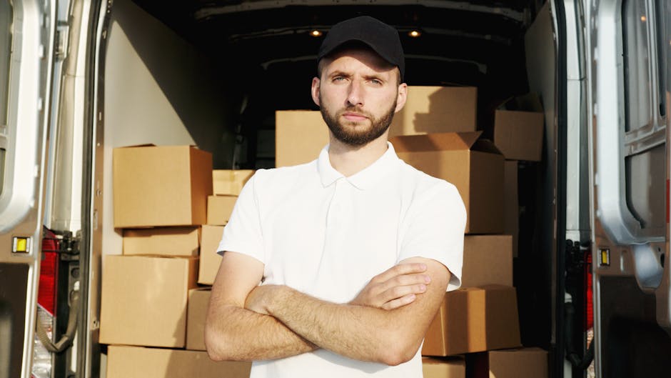 A man dressed in a blue jacket, dark trousers, and white sneakers is loading cardboard boxes into the back of a white commercial van parked on a cobblestone street in an urban setting. The van's rear 