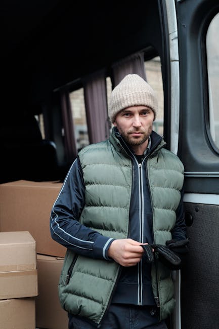 A man with a beard and wearing a black cap, white polo shirt, and jeans stands inside the open back of a delivery van near Gunnersbury Park, holding a cardboard box. The van is filled with several nea