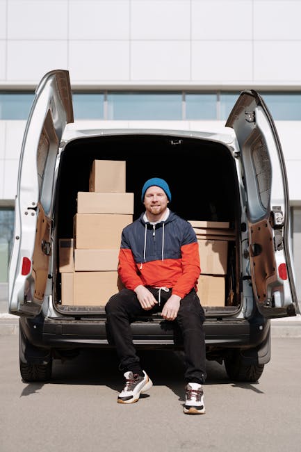 A man wearing a white t-shirt, jeans, and a black cap is loading three medium-sized cardboard boxes into the side of a white delivery van parked on a residential street. The van, branded with black an